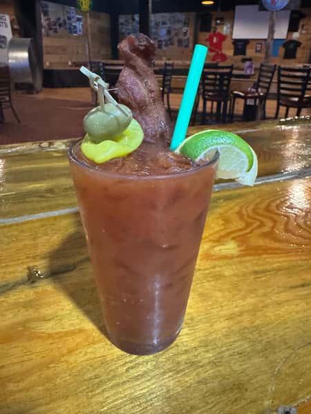 Bloody Mary garnished with bacon, green olives, a pepperoncini, and a lime wedge, served in a tall glass with a straw.