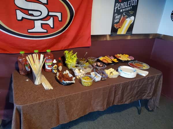 Self-serve Bloody Mary bar setup with tomato juice, sauces, skewers, garnishes, bacon, celery, cheese, and meats on a brown tablecloth.