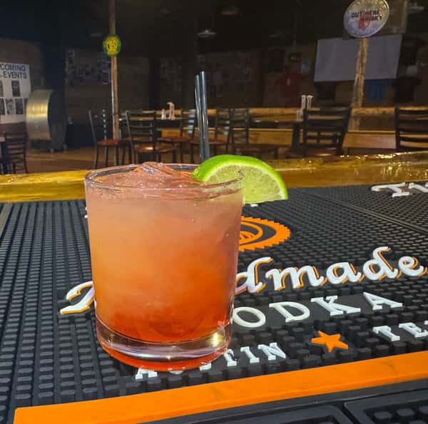 Red and orange gradient cocktail served on the bar with ice, a lime wedge garnish, and a black straw.