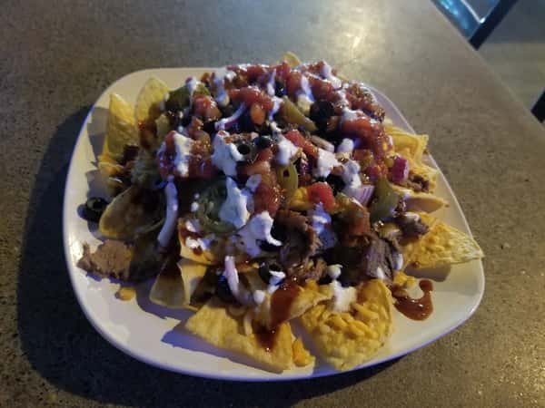 This image shows a hearty plate of nachos loaded with toppings including cheese, refried beans, shredded meat, jalapeños, black olives, diced tomatoes, onions, salsa, and a drizzle of sour cream or crema. It looks like a satisfying bar-style appetizer, likely from the same establishment shown in earlier images.