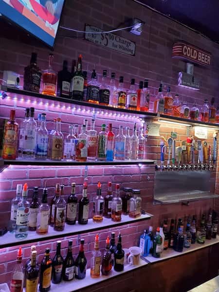 Well-stocked bar with a wide variety of liquor bottles, beer taps, and backlit shelves against a brick wall, featuring humorous signs and a TV overhead.