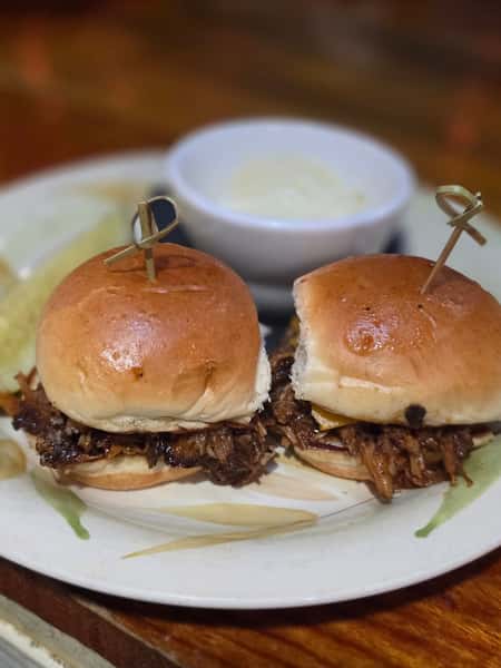 Two pulled pork sliders on soft buns served with chips and a side of creamy dipping sauce on a decorative plate.