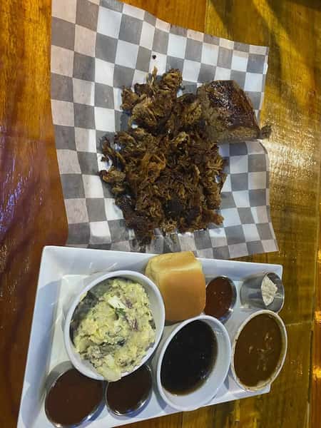 Pulled pork and brisket served on checkered paper with sides of potato salad, baked beans, dinner roll, and assorted barbecue sauces.
