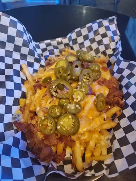 Cheese fries topped with crispy bacon and sliced jalapeños, served in a basket with black and white checkered paper.