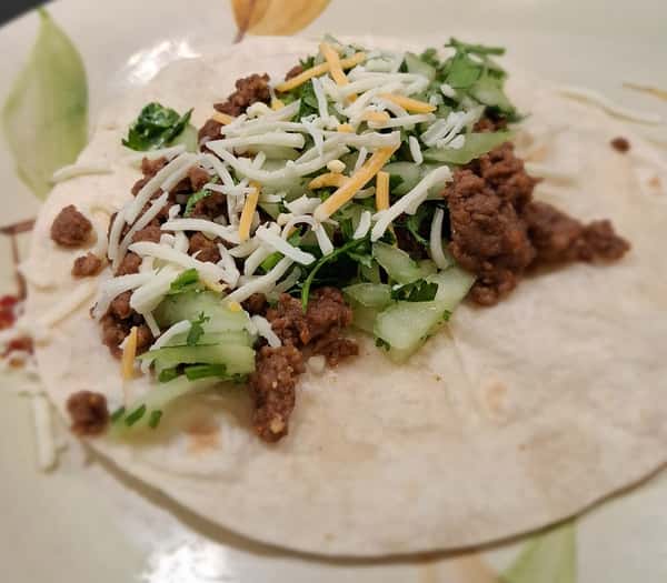Soft taco with seasoned ground beef, shredded cheese, cucumber slices, and fresh cilantro on a flour tortilla.