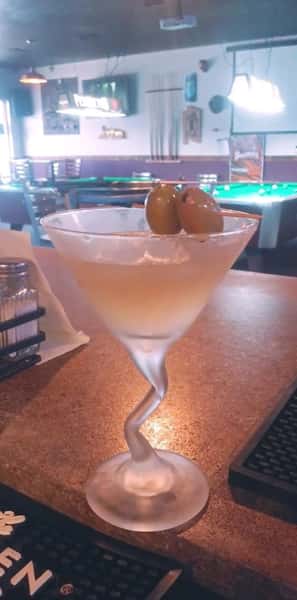 Chilled martini in a twisted stem glass garnished with two green olives, served on a bar counter near a pool table.