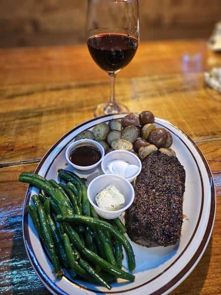 Grilled steak with seasoned green beans, roasted potatoes, sauces, and a glass of red wine served on a wooden table.