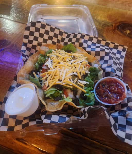 This is a taco salad served in a crispy tortilla bowl, topped with shredded cheese, lettuce, tomatoes, and black olives. It comes with sides of sour cream and salsa.