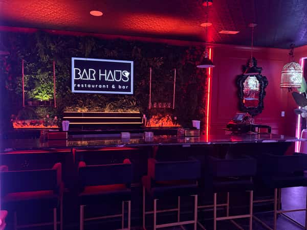 Moody neon-lit bar with velvet chairs, lush green wall, and a glowing "Bar Haus restaurant & bar" sign above the counter.