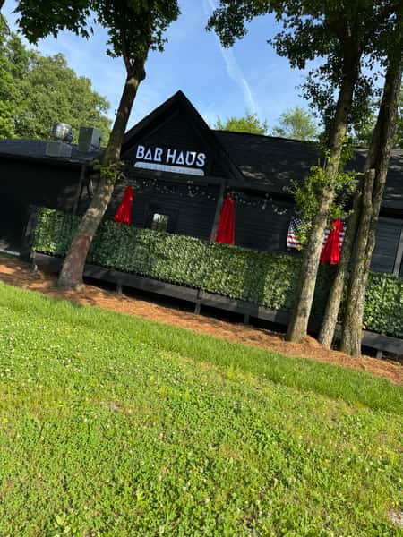 Exterior of Bar Haus, a dark-painted building with red umbrellas, greenery-covered patio railing, and trees in front on a sunny day.