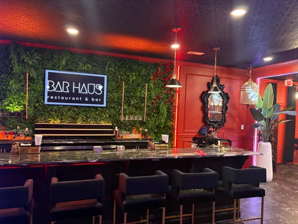 Chic bar with velvet stools, marble counter, lush green wall, neon "Bar Haus" sign, and moody red and black lighting.