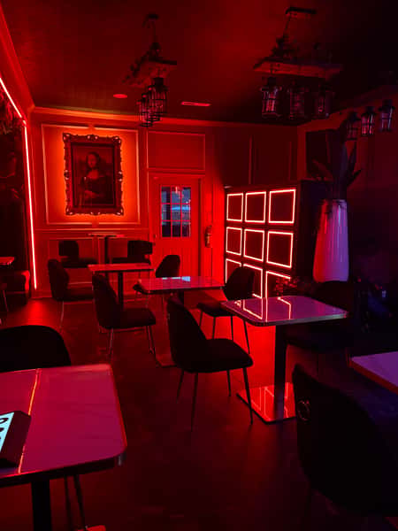 Moody red-lit lounge with marble-top tables, black chairs, a neon square wall display, and a framed Mona Lisa painting on the wall.