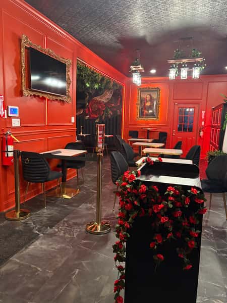 Interior of a restaurant with red walls, featuring a TV, vintage-style portraits, a flower-decorated counter, and elegant black chairs with gold accents.