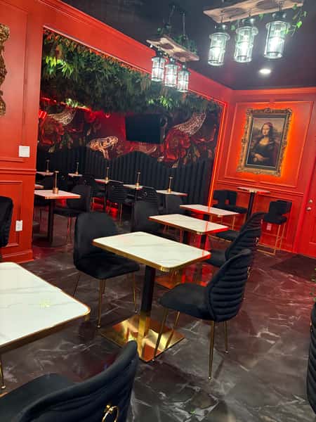 Interior of a restaurant with red walls, featuring a TV, vintage-style portraits, a flower-decorated counter, and elegant black chairs with gold accents.