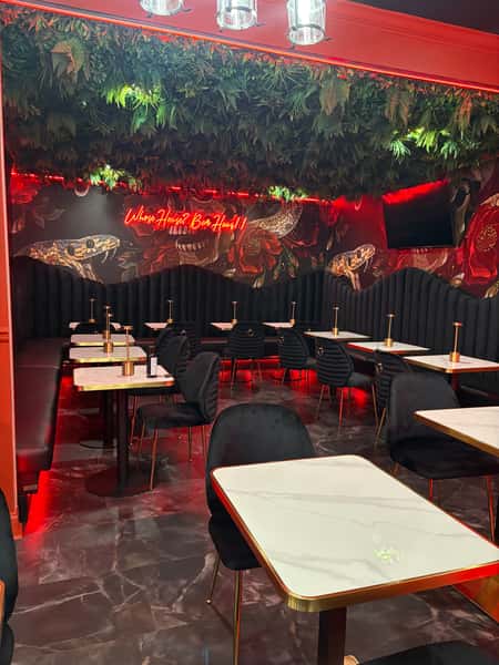 Stylish dining area with black velvet chairs, marble-top tables, lush greenery on the ceiling, and red neon sign reading "Whose House? Bar Haus!"