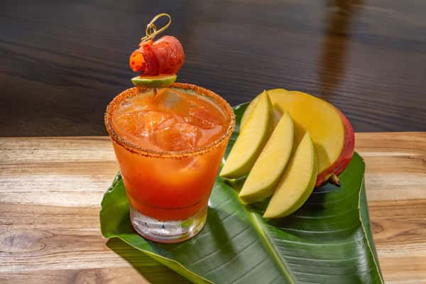 Spicy mango cocktail with a chili-salt rim, garnished with a cherry tomato skewer, served alongside fresh mango slices on a banana leaf