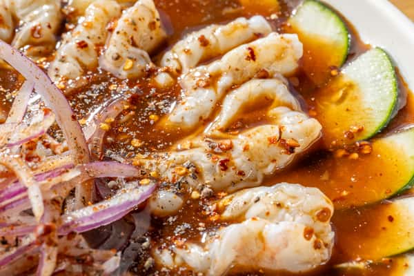 "Aguachile" Close-up of plump shrimp in a black salsa with red onion, cucumber slices, and spices