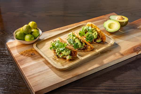 Three street-style tacos loaded with octopus, guacamole and fresh herbs on a wooden board, served alongside halved avocados and lime