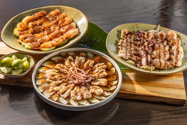 three-plate seafood spread featuring aguachile on half-moon sliced cucumber, scallops, and shrimp drizzled in sauce arranged on a wooden board with banana leaf