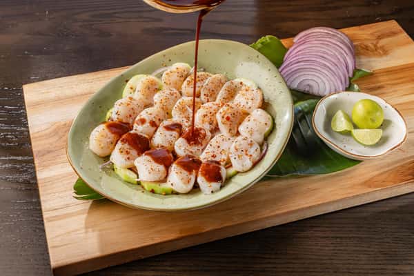 "Botana Callo De Hacha" Plump scallops arranged on a green plate, drizzled with dark sauce and dusted with chili powder, served with cucumber slices on a banana leaf