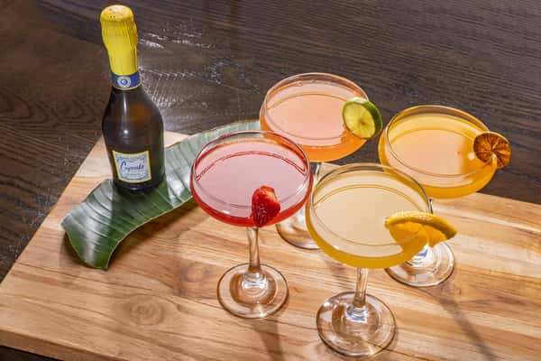 four cocktails in pink, red, and orange, and yellow garnished with fresh fruit, alongside a Cupcake prosecco bottle on a dark wood table