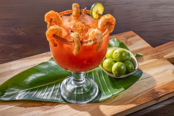 Large spicy tomato-based cocktail in a chili-rimmed glass, topped with dried shrimp and cucumber, served with limes on a banana leaf