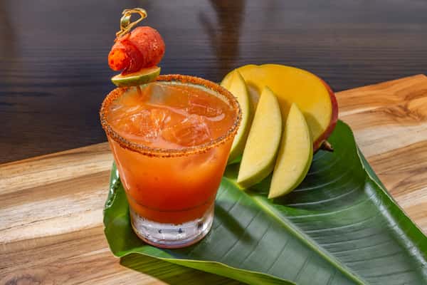 picy mango cocktail in a chili-rimmed glass garnished with a cherry tomato skewer, served alongside fresh mango slices on a banana leaf