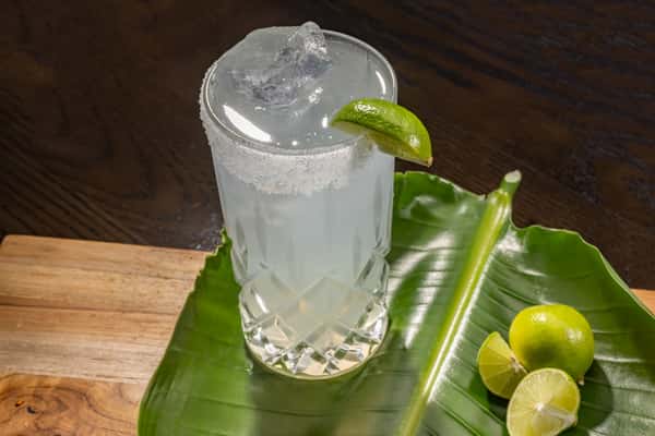 Iced clear citrus drink garnished with a lime wedge, served on a banana leaf alongside fresh limes on a wooden surface