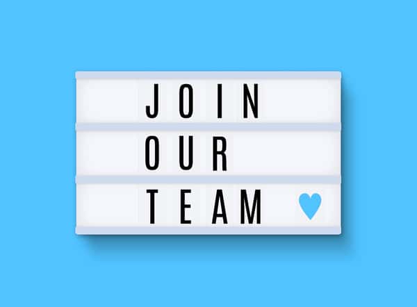 Light box sign reading 'Join Our Team' with a blue heart, displayed on a bright blue geometric background