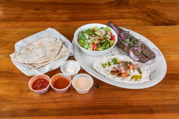 Grilled kafta kabob plate with rice, salad, pita bread, vegetables, and dipping sauces.