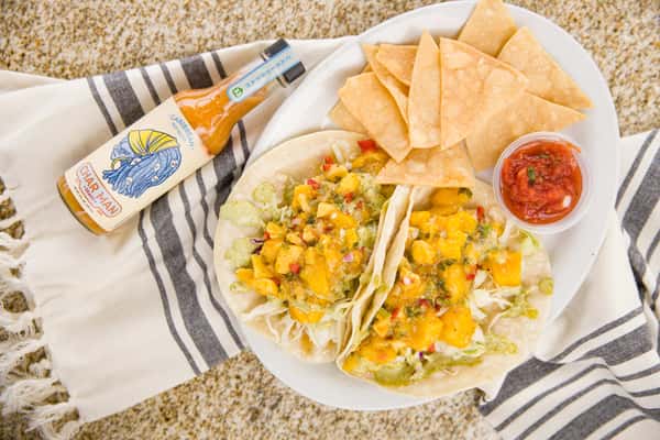 A vibrant plate of fish tacos filled with crispy battered fish, topped with fresh corn salsa and diced vegetables. The tacos are served with a side of tortilla chips and a small portion of salsa, creating a colorful and flavorful coastal-inspired dish.