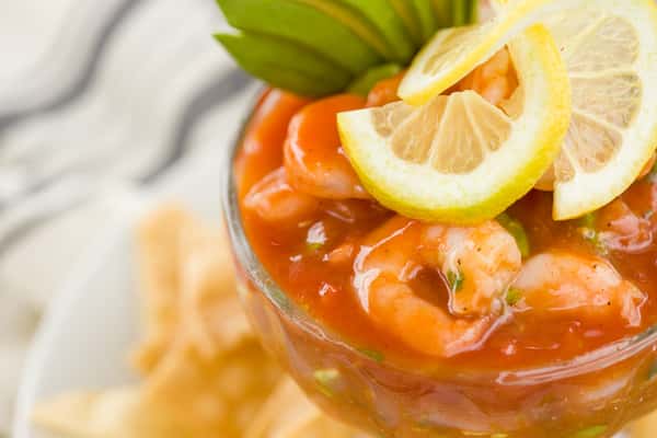 A close-up of a refreshing seafood cocktail served in a glass, filled with shrimp, a rich tomato-based sauce, and garnished with fresh lime slices. Crispy tortilla chips sit alongside, adding texture and a casual coastal feel.