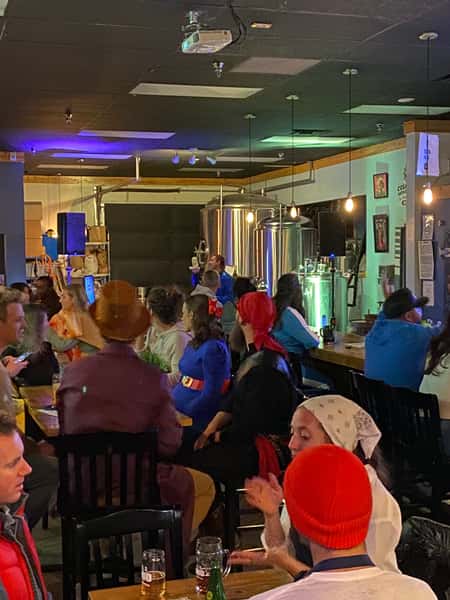Patrons in colorful costumes gathered at tables and the bar, enjoying drinks and entertainment in the brewery's taproom.
