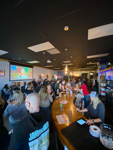 Guests enjoying drinks and conversation at a lively bar inside Old 121 Brewhouse, with a large screen showing colorful visuals in the background.