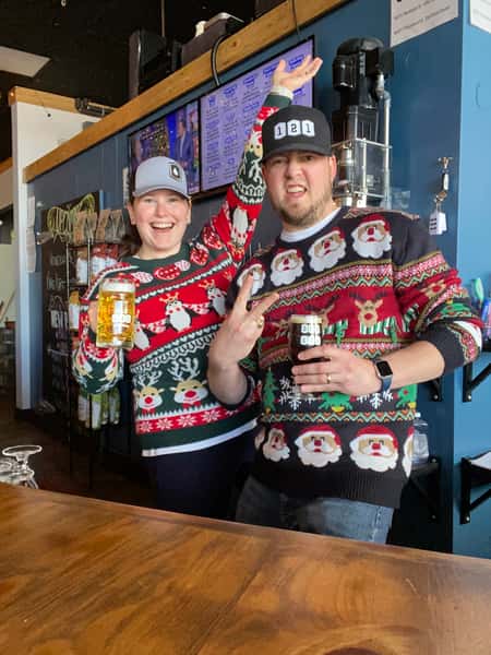 Two smiling people wearing festive Christmas sweaters and brewery hats, holding beer mugs inside a taproom.
