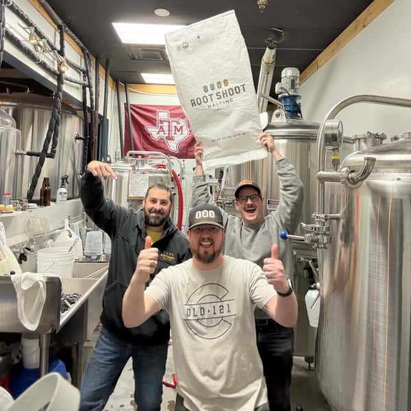 Three brewers posing in the brewing area, holding a large Root Shoot Malting grain bag and giving thumbs up.