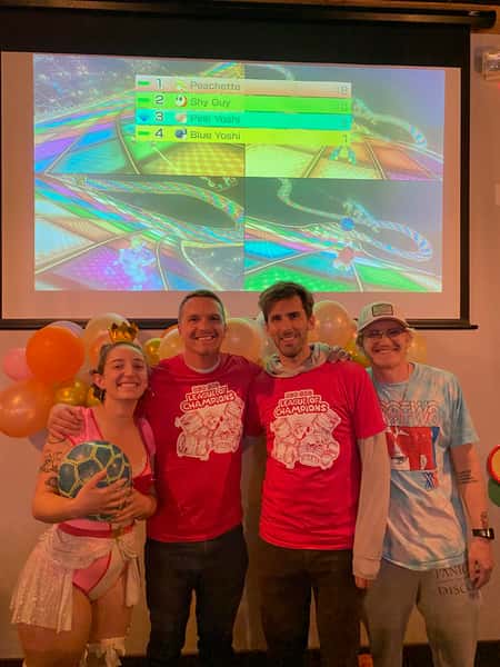 Four smiling contestants posing in front of a Mario Kart game screen, wearing matching Old 121 League of Champions shirts.