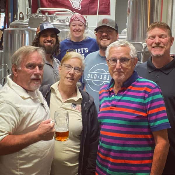 Smiling group of guests posing in front of stainless steel brewing tanks at Old 121 Brewhouse, with one person holding a pint of amber beer.