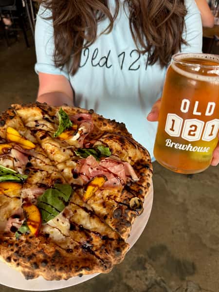Freshly baked artisan pizza topped with prosciutto, basil, peaches, and balsamic glaze, paired with a pint of golden beer in an Old 121 Brewhouse glass.