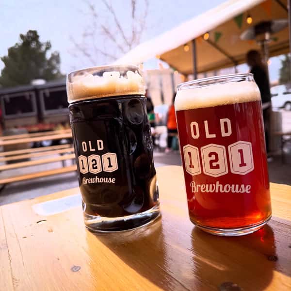 Close-up of a dark beer stein and amber pint glass with Old 121 Brewhouse logos, sitting on an outdoor picnic table.