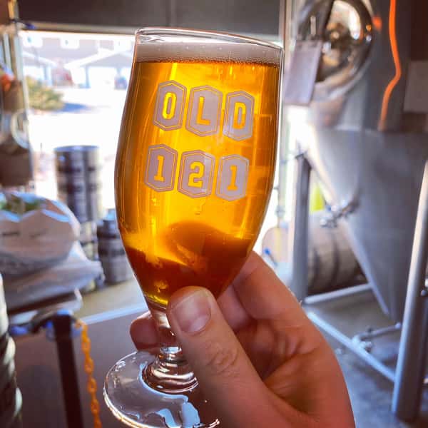 Close-up of a tulip glass filled with golden beer, held up in front of brewing tanks inside Old 121 Brewhouse.