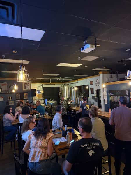 Guests seated at tables and the bar, enjoying craft beer and conversation while a trivia host speaks near the brewing tanks.
