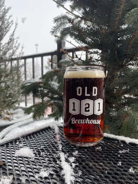 Pint of dark beer in an Old 121 Brewhouse glass sitting on a snow-dusted metal table with evergreen trees in the background.