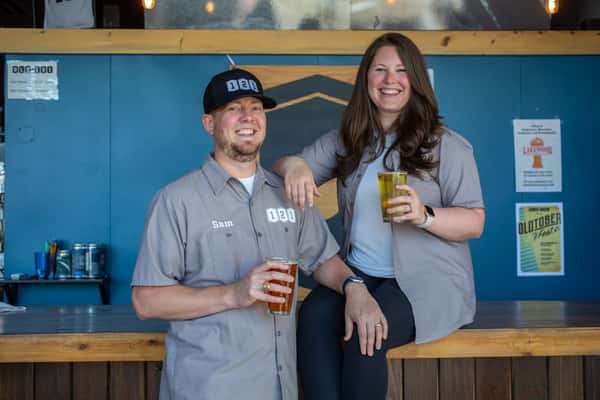 Two smiling brewery owners holding pints of beer while posing behind the bar at Old 121 Brewhouse, wearing branded shirts and hats.