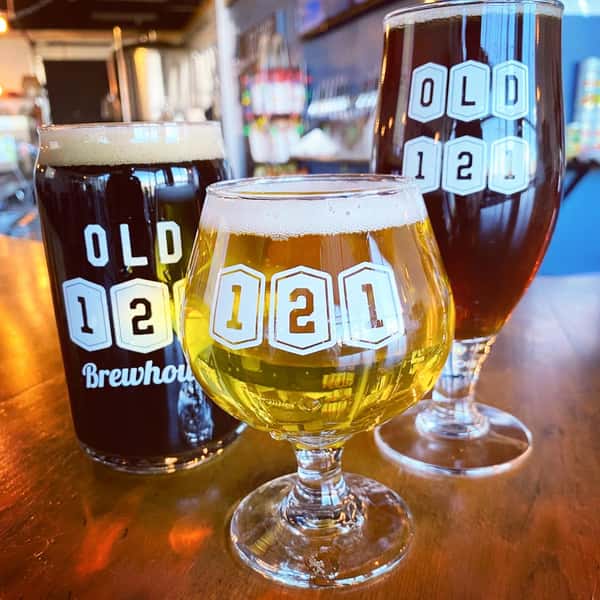 Close-up of three beers in branded Old 121 Brewhouse glasses, showcasing a golden ale, dark stout, and rich amber brew on a wooden bar.