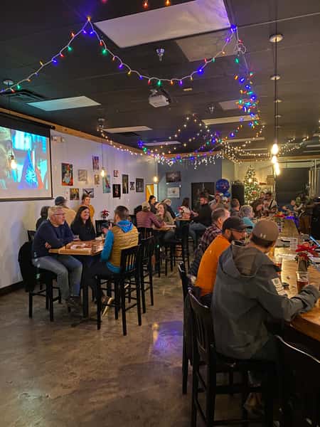 Guests enjoying drinks and conversation inside Old 121 Brewhouse, under colorful string lights and holiday decorations, with a large movie screen in the background.