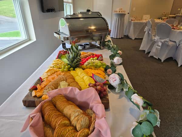 A beautifully arranged catering spread featuring a colorful assortment of fruits, cheeses, and crackers, with a decorative pineapple centerpiece and elegant table settings in the background.