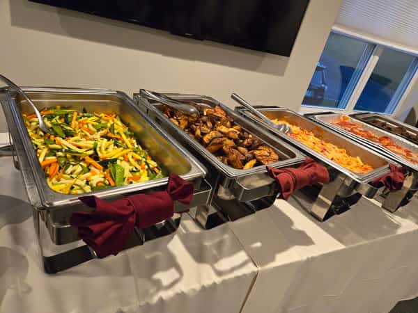 A catering setup featuring three chafing dishes with colorful mixed vegetables, grilled chicken, and pasta, accompanied by neatly folded maroon napkins on a white tablecloth.