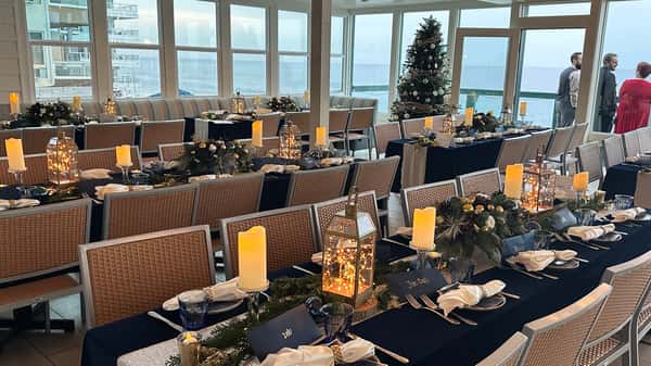 Elegant indoor oceanfront dining setup at Driftwood Kitchen in Laguna Beach, featuring navy blue tablecloths, glowing lanterns, holiday greenery, and candle centerpieces with a decorated Christmas tree in the background and panoramic Pacific Ocean views.