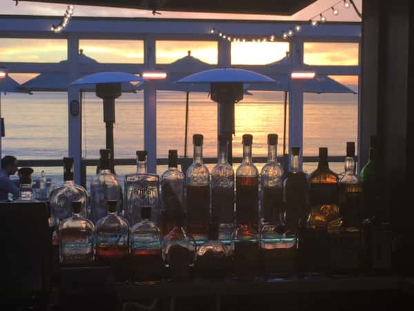 Backlit liquor bottles on a beachside bar at The Deck on Laguna Beach, silhouetted against a glowing Pacific Ocean sunset with string lights and patio heaters reflecting a warm, coastal ambiance.